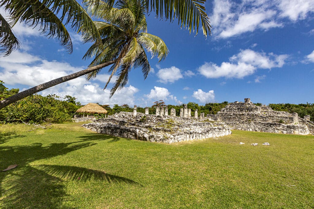 Ancient Mayan ruins in Cancun with stone columns and structures under a bright, sunny sky with scattered clouds. A tall palm tree leans over the grassy area, casting a shadow on the ground. Lush vegetation can be seen in the background.