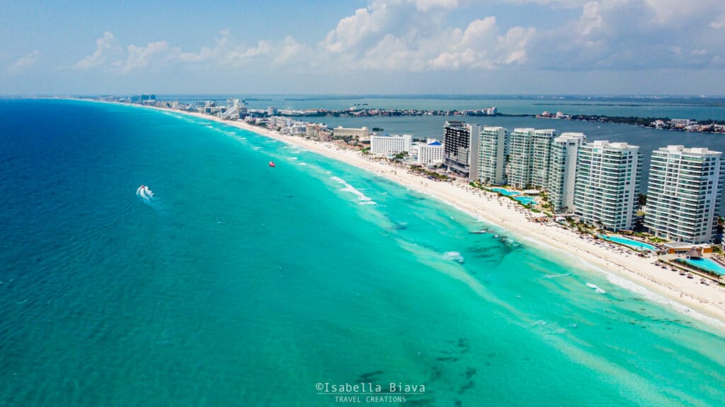 Aerial view of a coastal city with high-rise buildings along a sandy beach and turquoise ocean, stretching towards the horizon under a blue sky with scattered clouds—perfect for Cancun activities for couples looking to enjoy stunning scenery together.