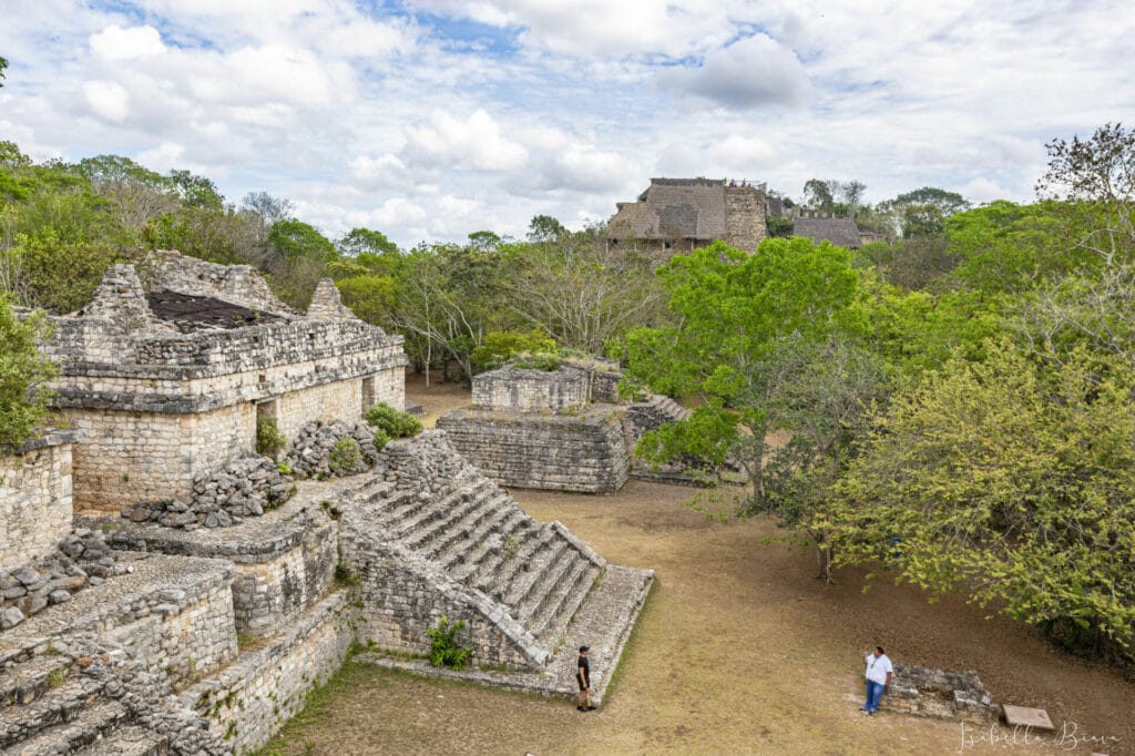Stone ruins of Ek' Balam, an ancient Mayan city with several pyramids and structures, surrounded by lush greenery. Two people are seen walking in the foreground, exploring the breathtaking Mayan ruins in Cancun.